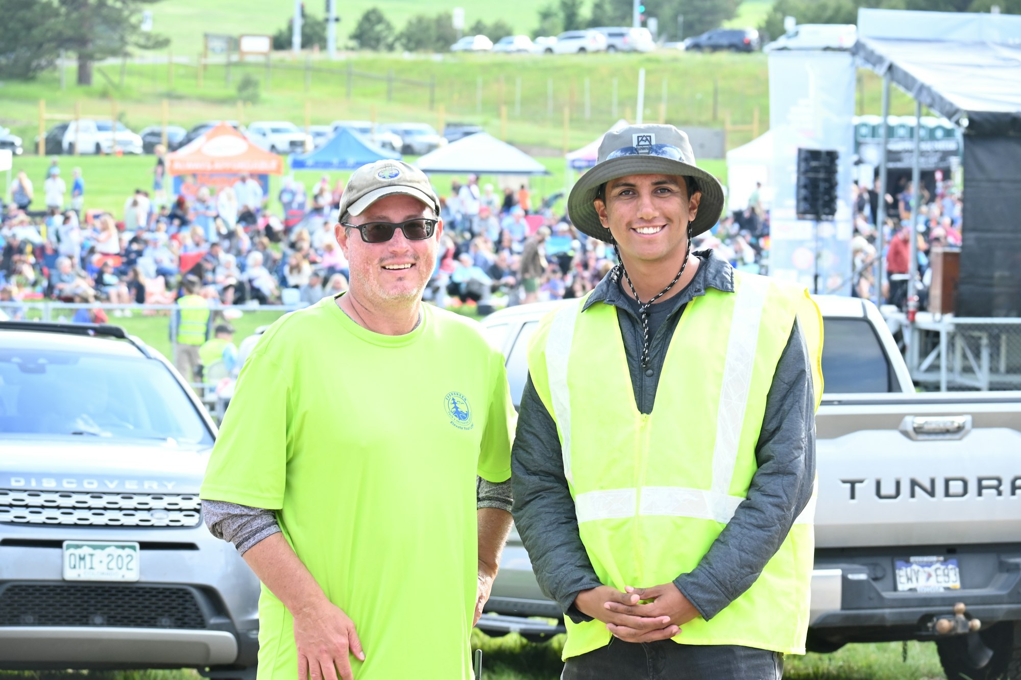Two people in bright safety gear smile at an outdoor event with a crowd and trucks in the background.