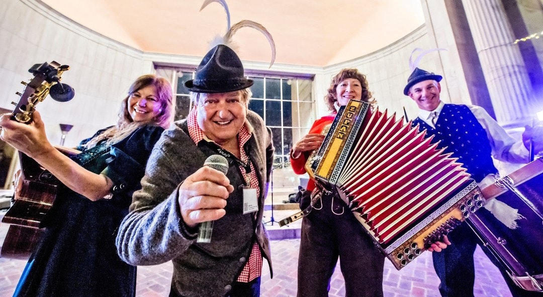 Four musicians, cheerfully posing with instruments: guitar, microphone, accordion, and contrabass, wearing Bavarian-style hats and attire.
