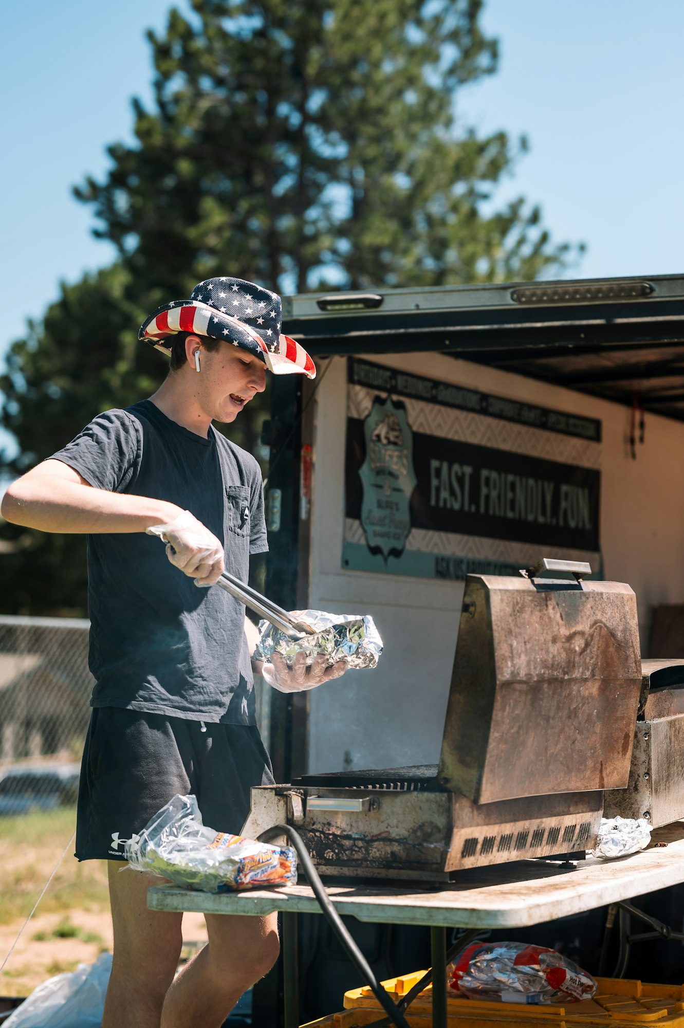 Person grilling outside, wearing a patriotic cowboy hat, listening to earbuds.