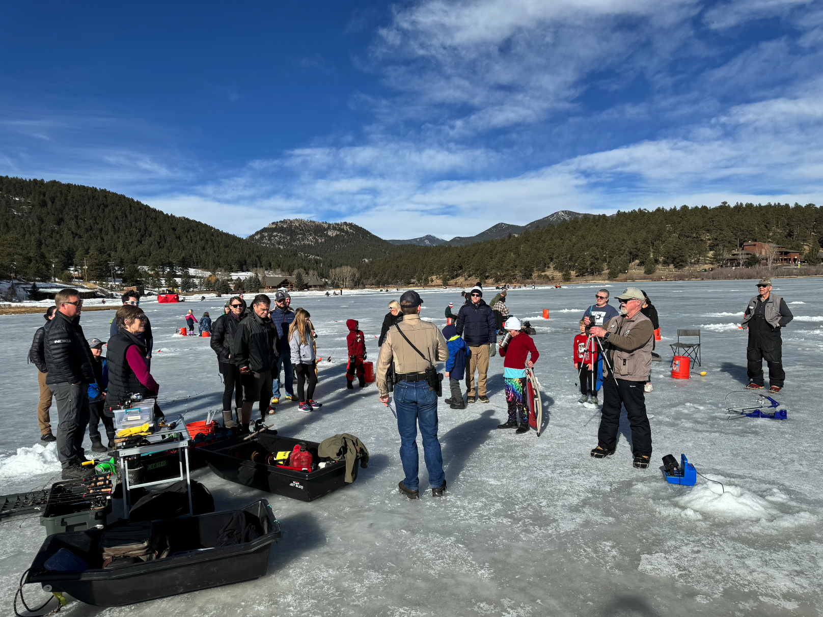 A group of people on a frozen lake, possibly ice fishing, surrounded by gear and scenic snowy mountains in the background.