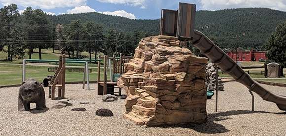 A playground with a rock climbing structure and slide, surrounded by trees under a blue sky.