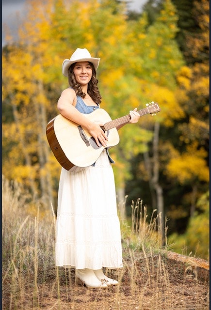 A woman in a white dress and cowboy hat stands outdoors holding an acoustic guitar, surrounded by autumn foliage.