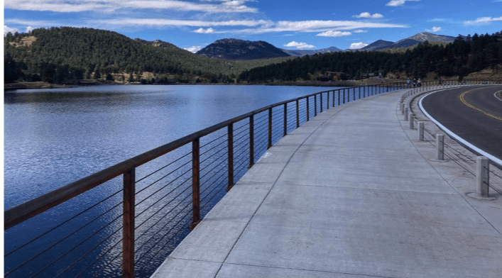 A lakeside walkway with a railing leading towards a mountainous backdrop under a partly cloudy sky.