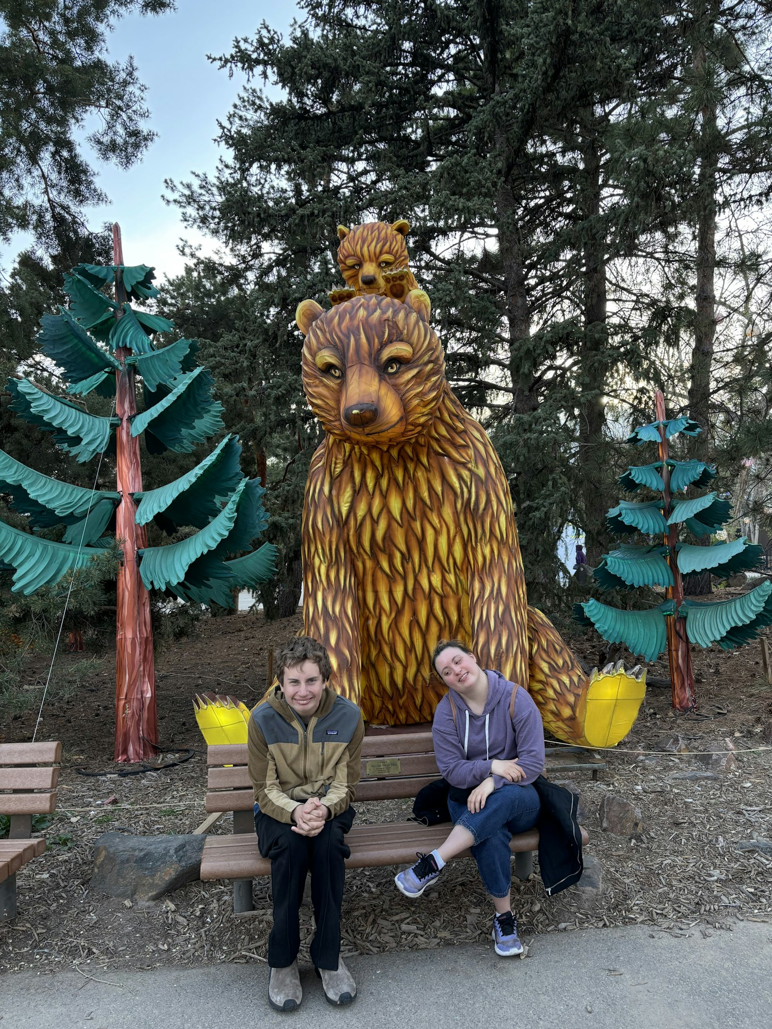 Two people sitting on a bench with a large bear statue behind them, surrounded by trees and foliage.