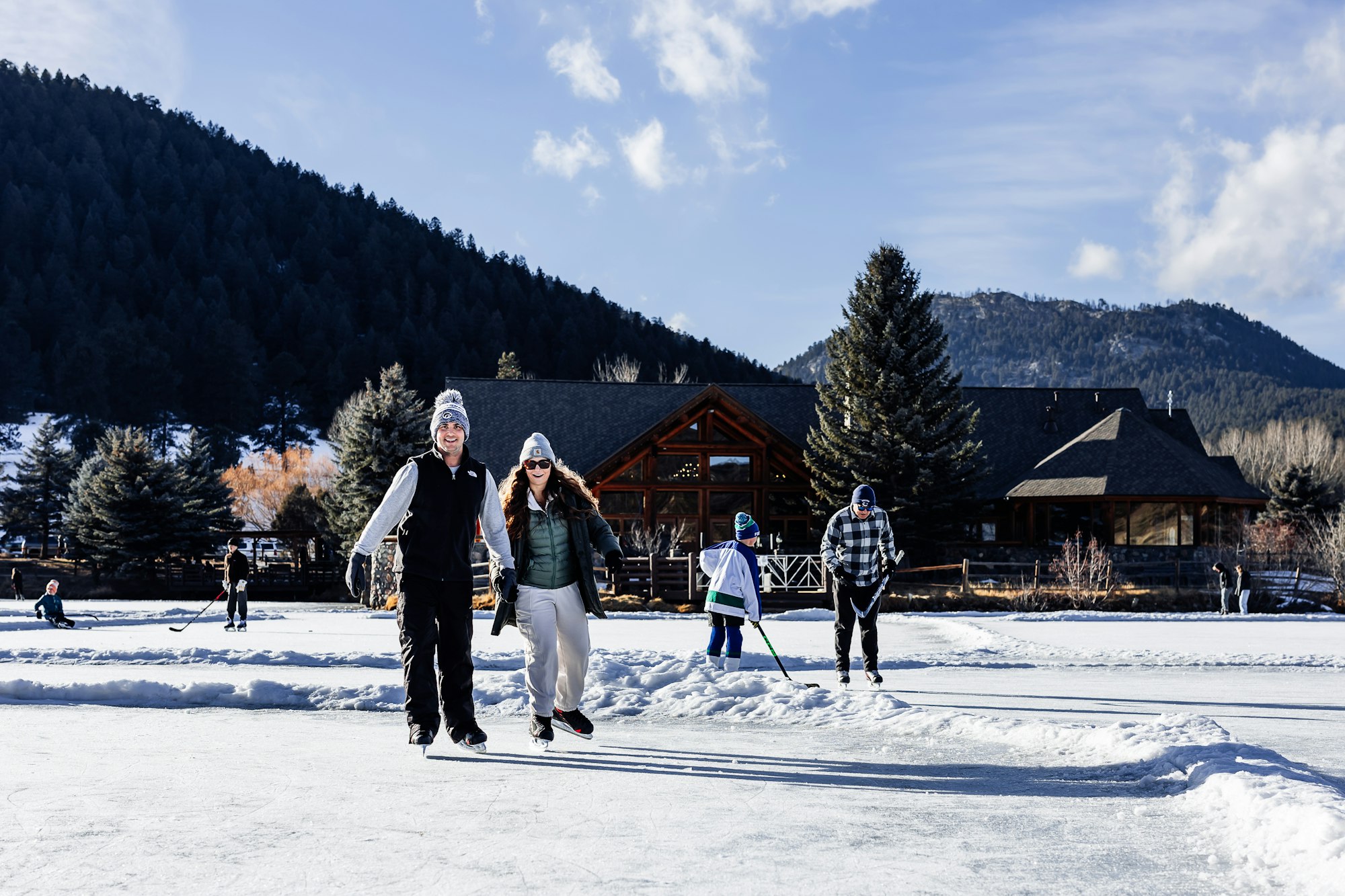 People ice skating outdoors near a wooden cabin with snowy mountains and pine trees in the background.