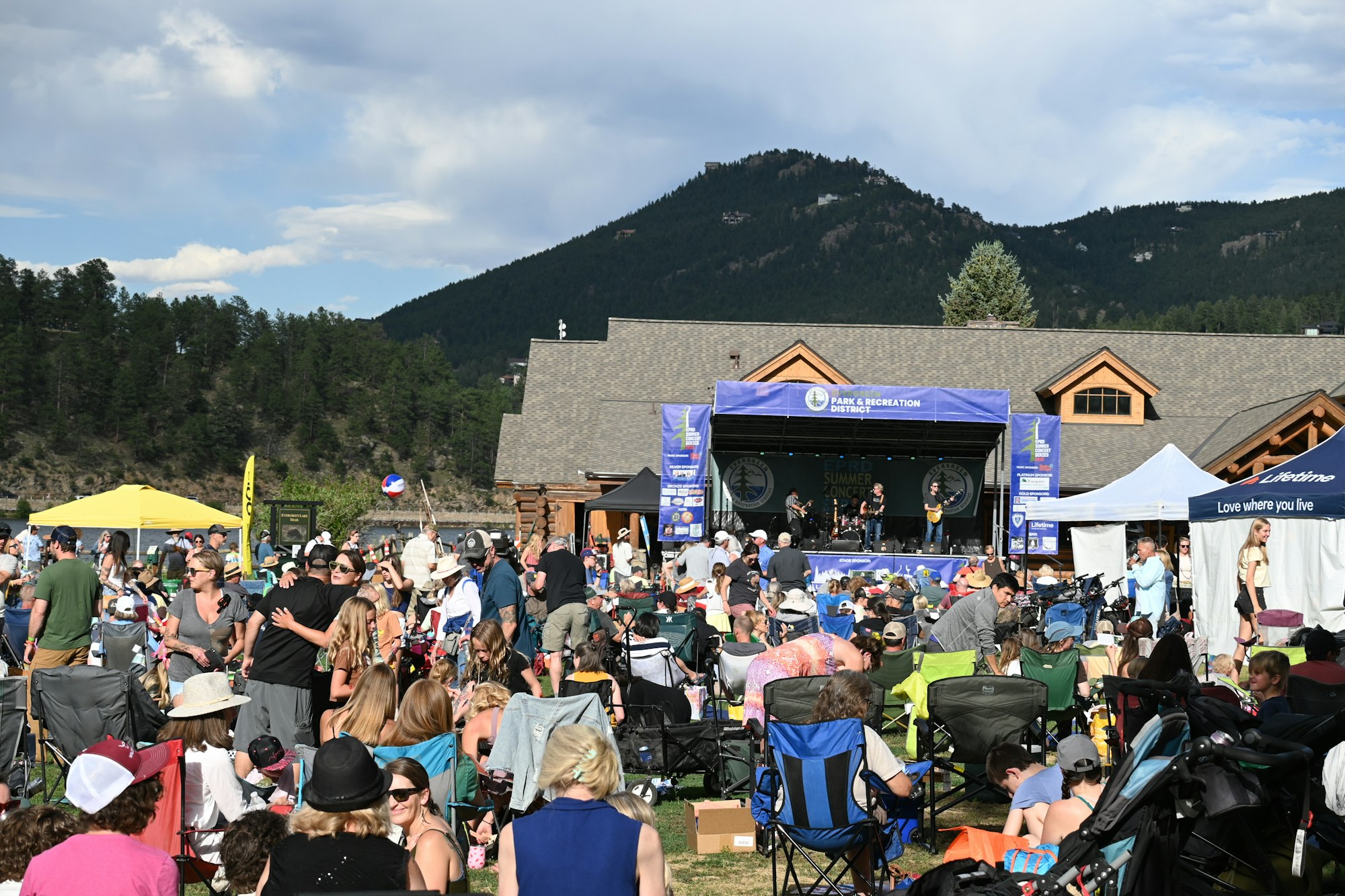 Outdoor concert with a stage, band, and crowd on lawn chairs. Mountain backdrop and sunny weather.