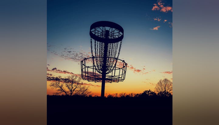 Disc golf basket silhouette against a sunset sky.