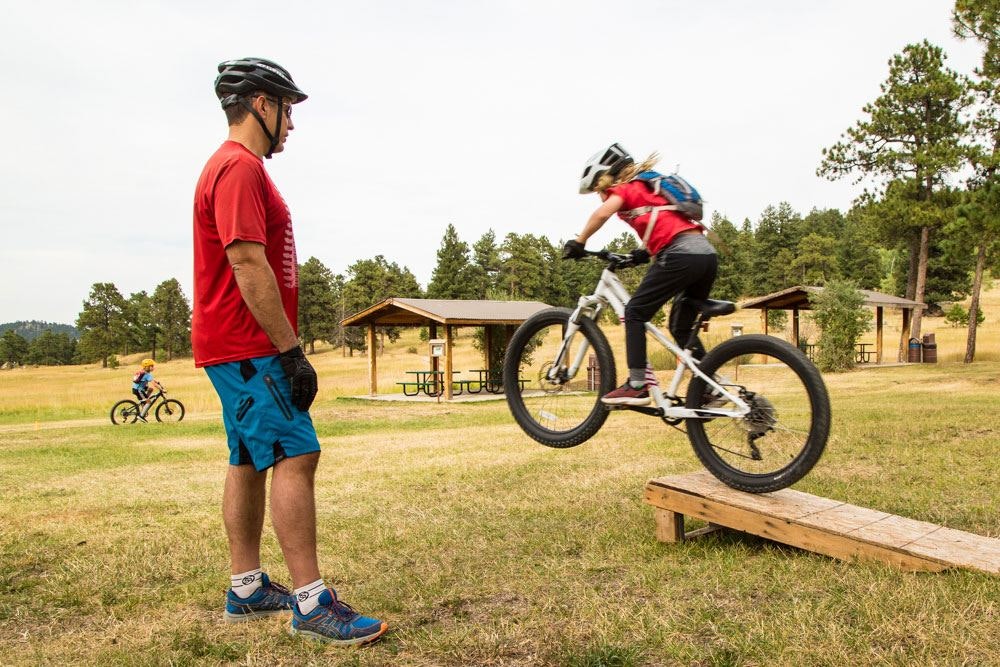 A person watches as another jumps a mountain bike off a ramp in a park setting.