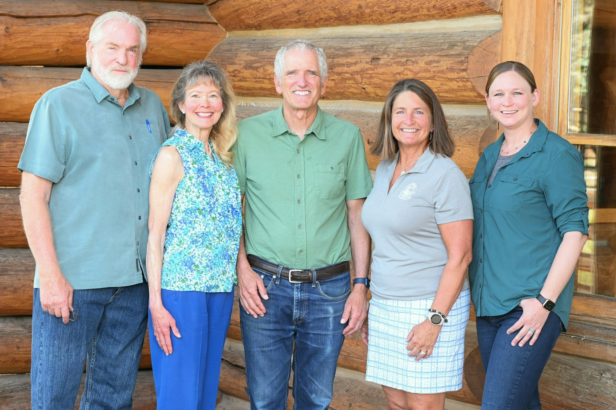 A group of five people stands together in front of a wooden cabin, smiling and dressed casually.