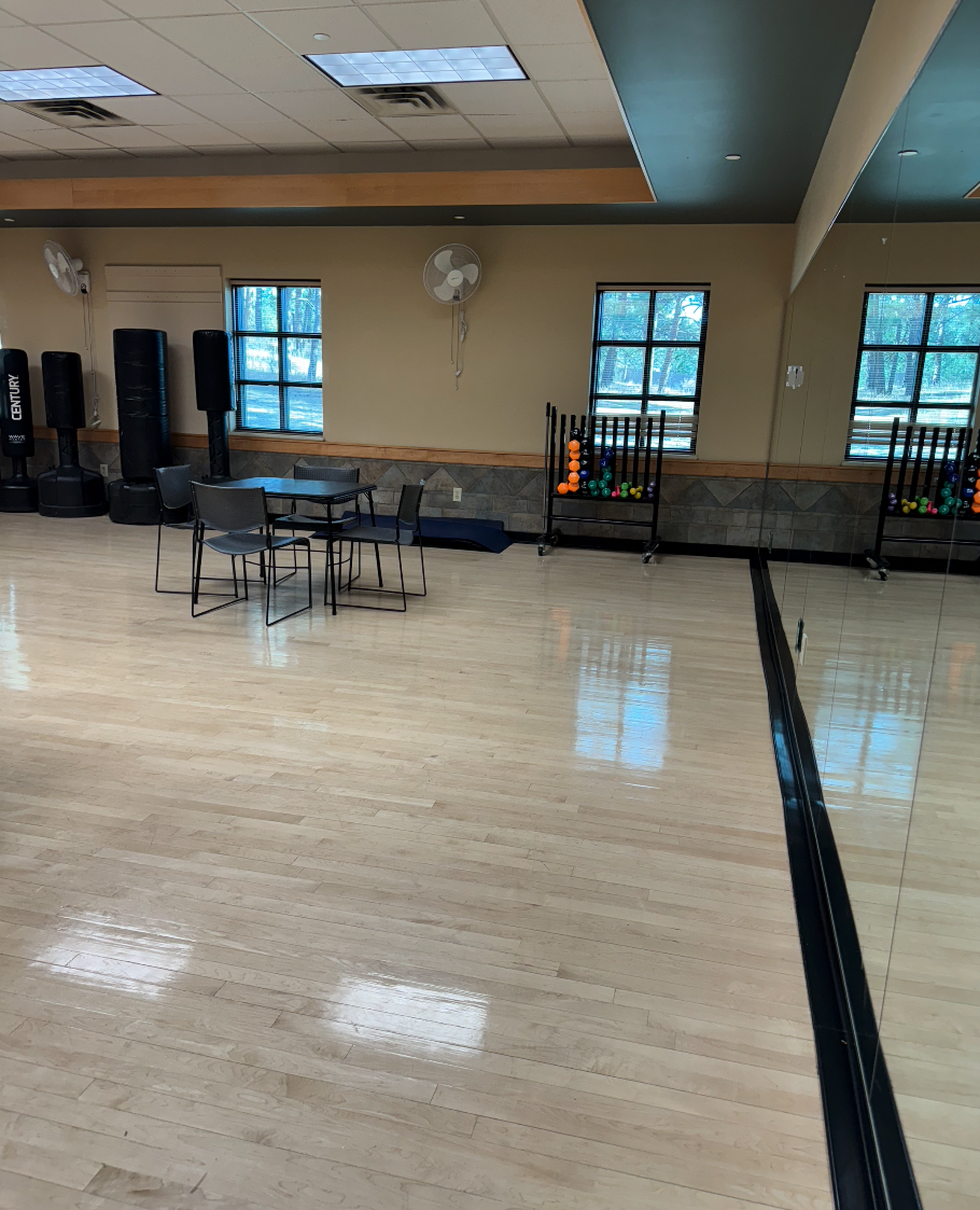 A gym room with mirrors, chairs, tables, punching bags, exercise balls, and large windows.