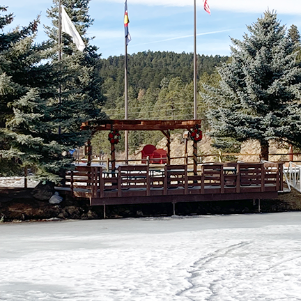 A frozen pond in front of a wooden gazebo with flags, surrounded by snow-dusted trees under a blue sky.