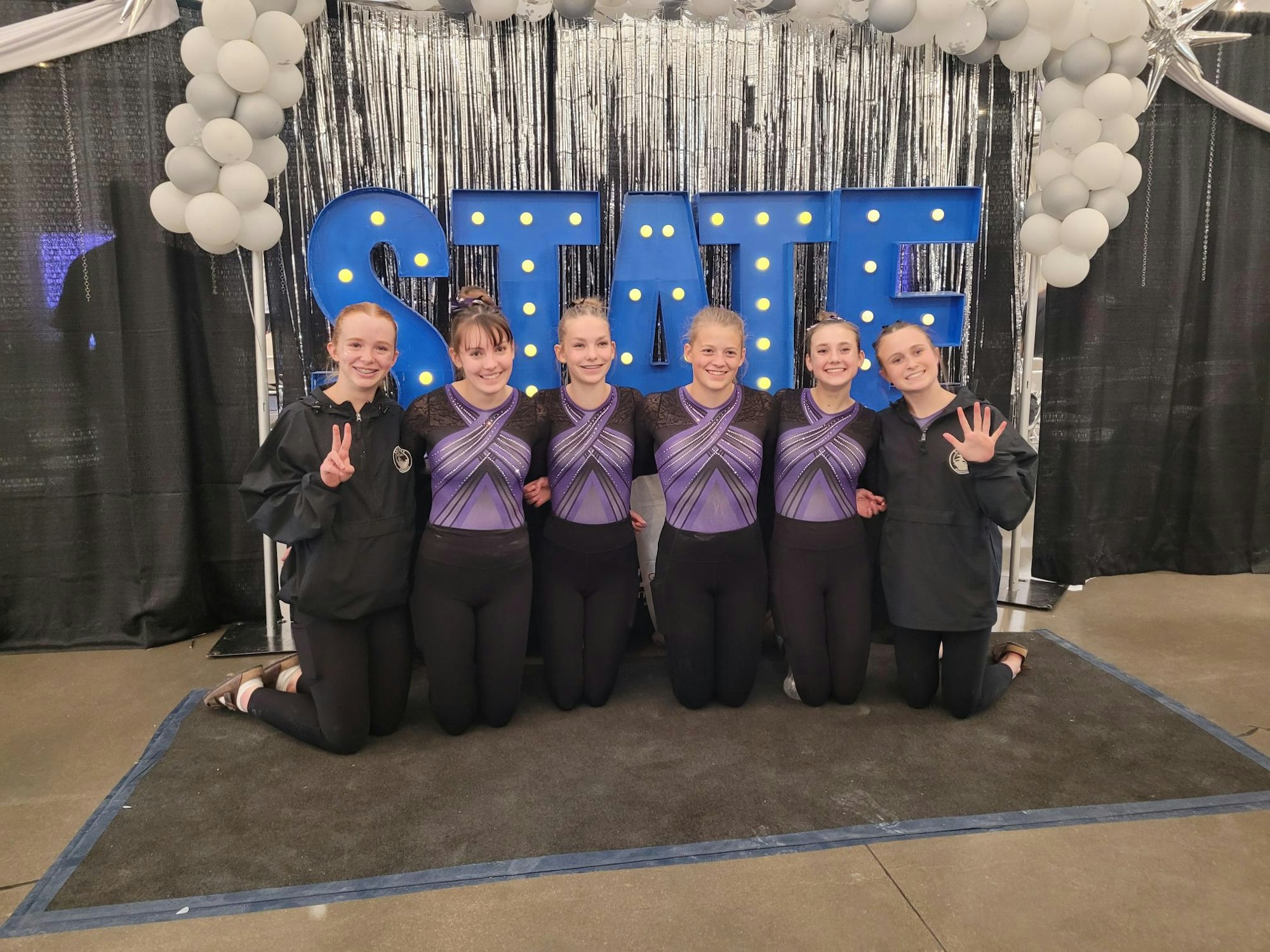 Six people posing in athletic attire with a "STATE" backdrop and balloons.