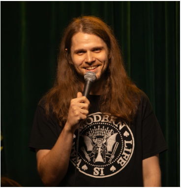 A person with long hair is holding a microphone and smiling, standing on stage in front of a dark green curtain, wearing a black graphic t-shirt.