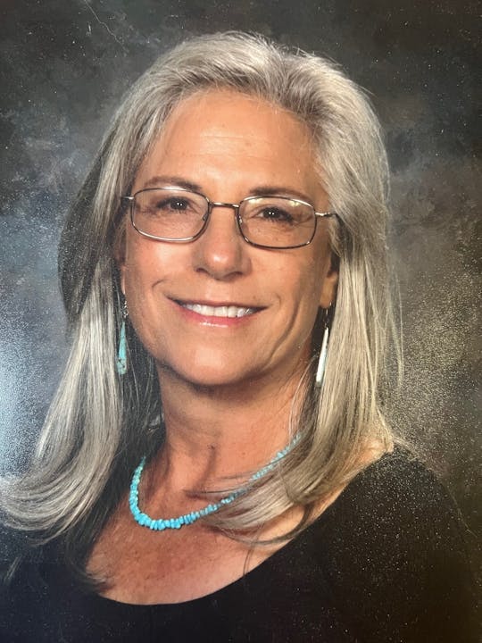 A woman with long gray hair, wearing glasses and turquoise jewelry, smiles for the camera against a dark background.
