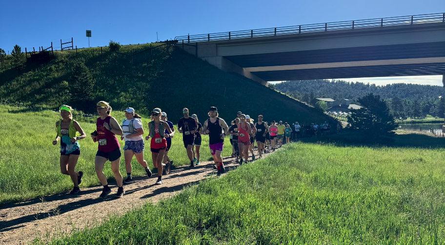 A group of runners on a sunny trail under a bridge, surrounded by green grass and trees.