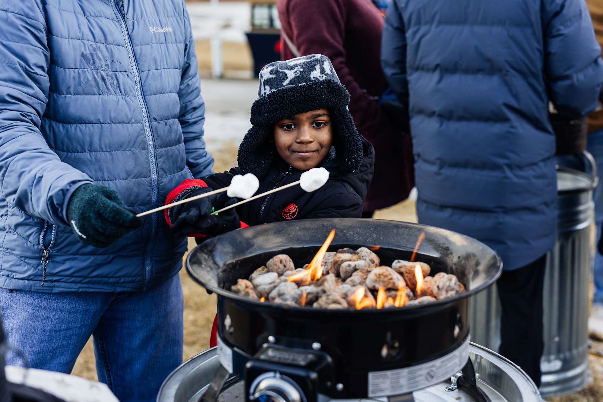 A child with marshmallows on sticks stands by a fire pit, enjoying a cozy moment outdoors with others in winter attire.