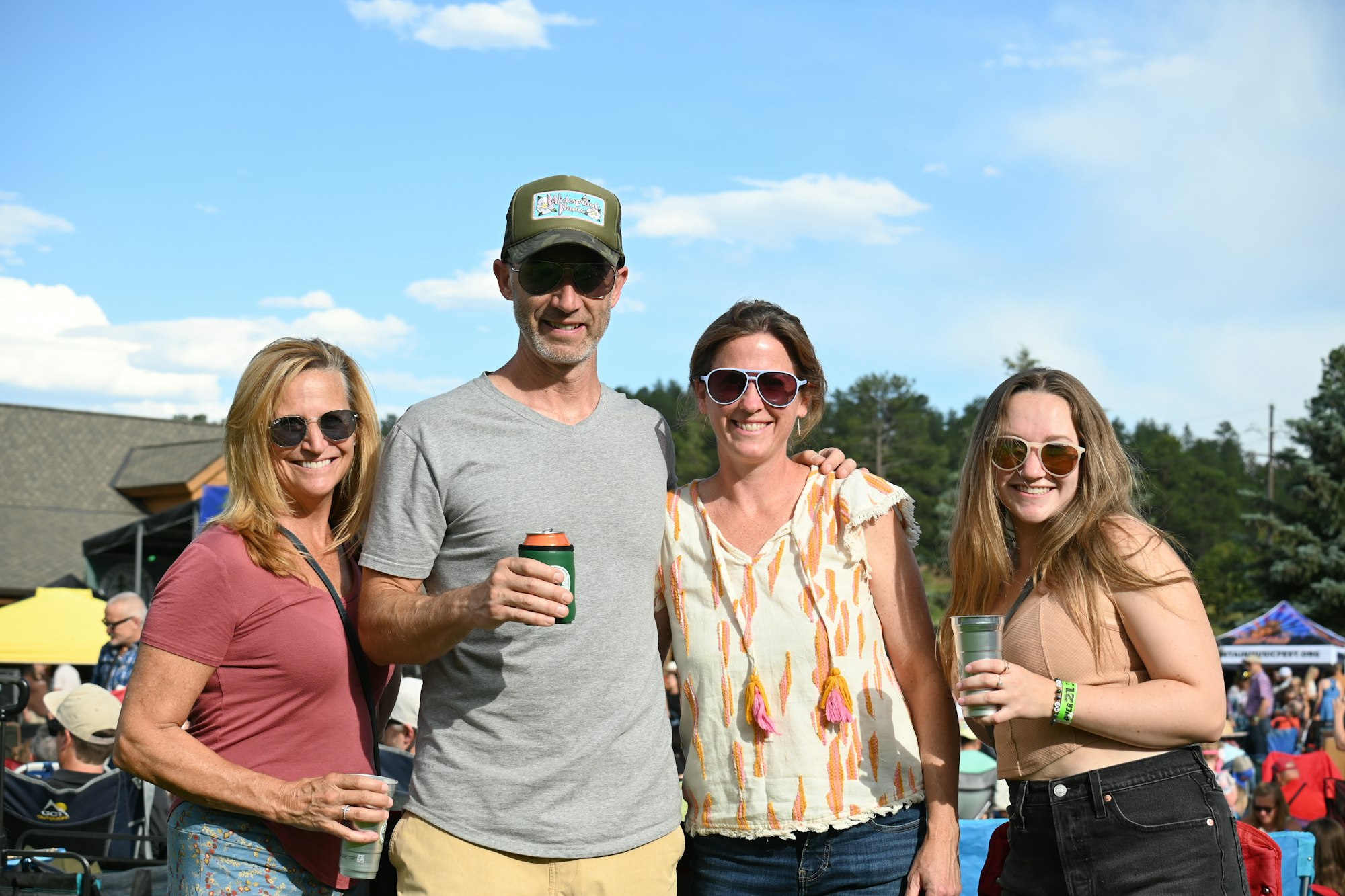 Four people wearing sunglasses, smiling, outdoors with drinks in hand.