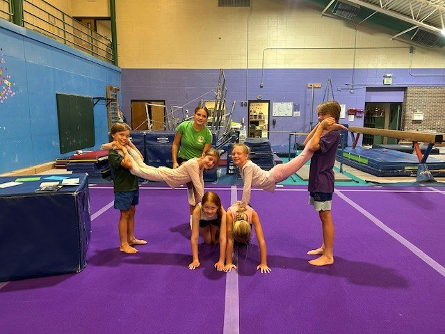 Children are practicing gymnastics poses on a purple mat, supported by each other, under the guidance of an instructor.