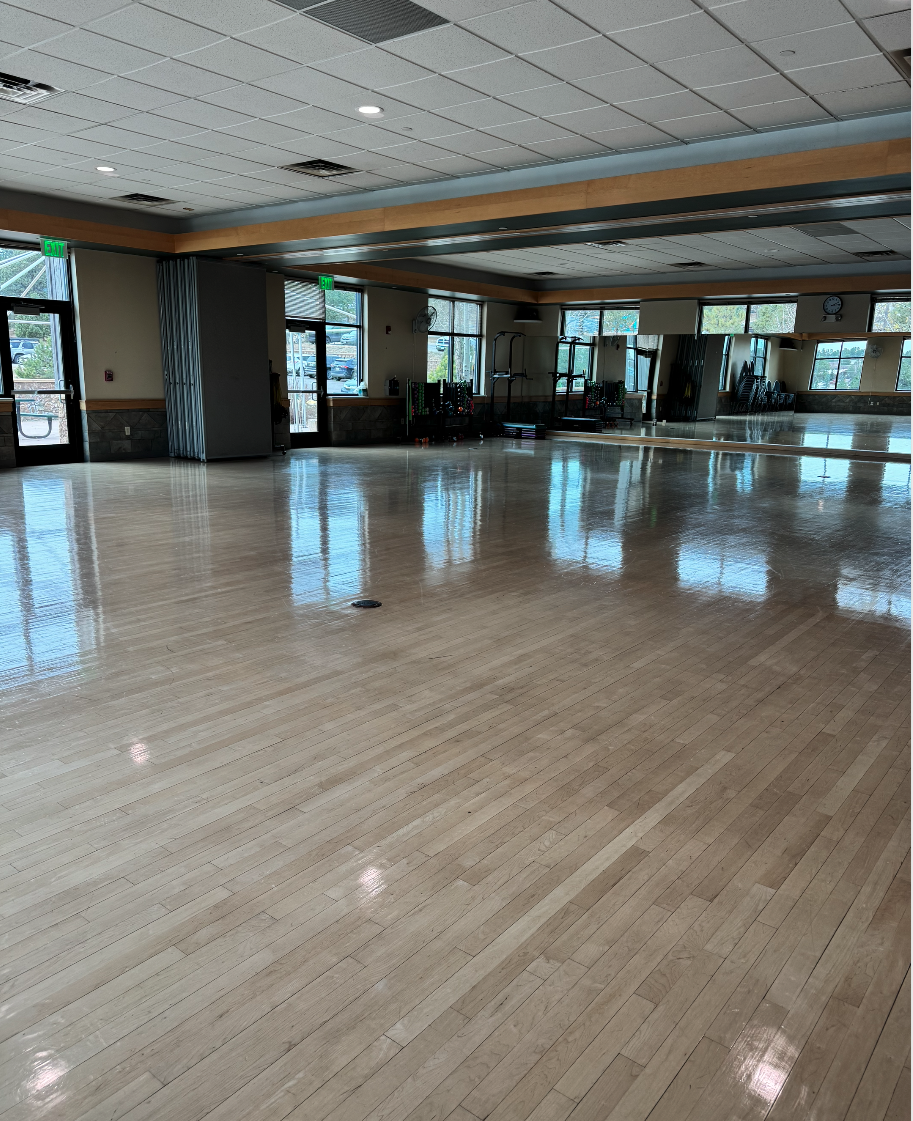 An empty indoor room with a wooden floor, mirrors, and exercise equipment near the windows.