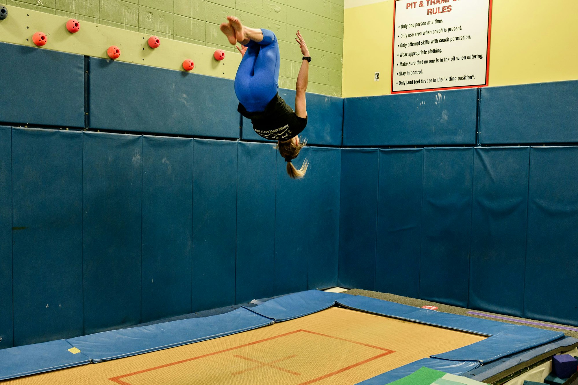 A person mid-flip above a gymnastics mat, with "Pit & Trampoline Rules" sign on the wall.