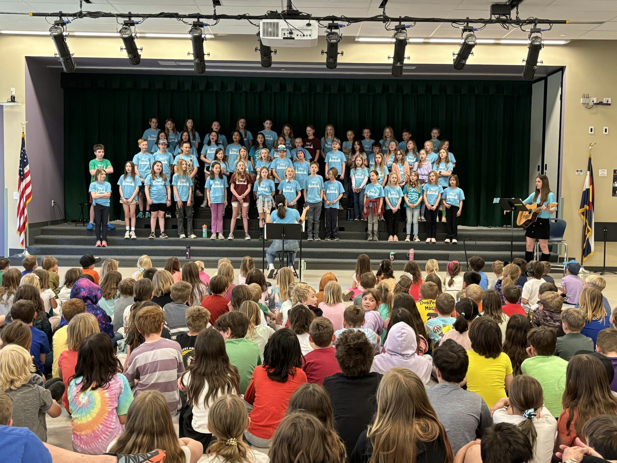 A choir of children wearing blue shirts performs on stage in front of an audience, with a person playing guitar to the side.