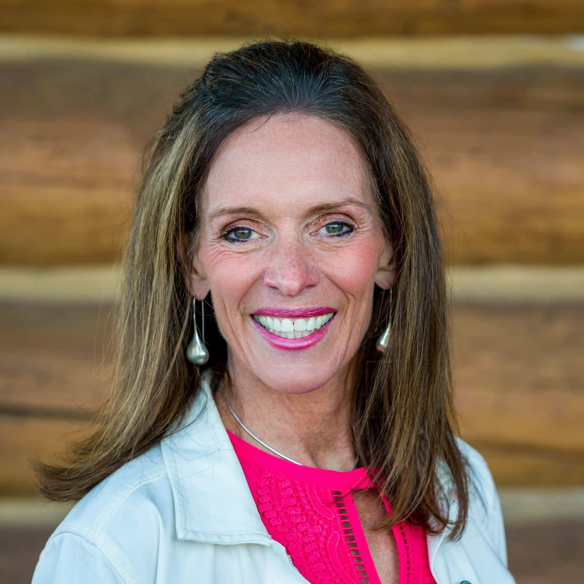 A smiling woman with long hair, wearing earrings, a pink top, and a white jacket against a wooden background.