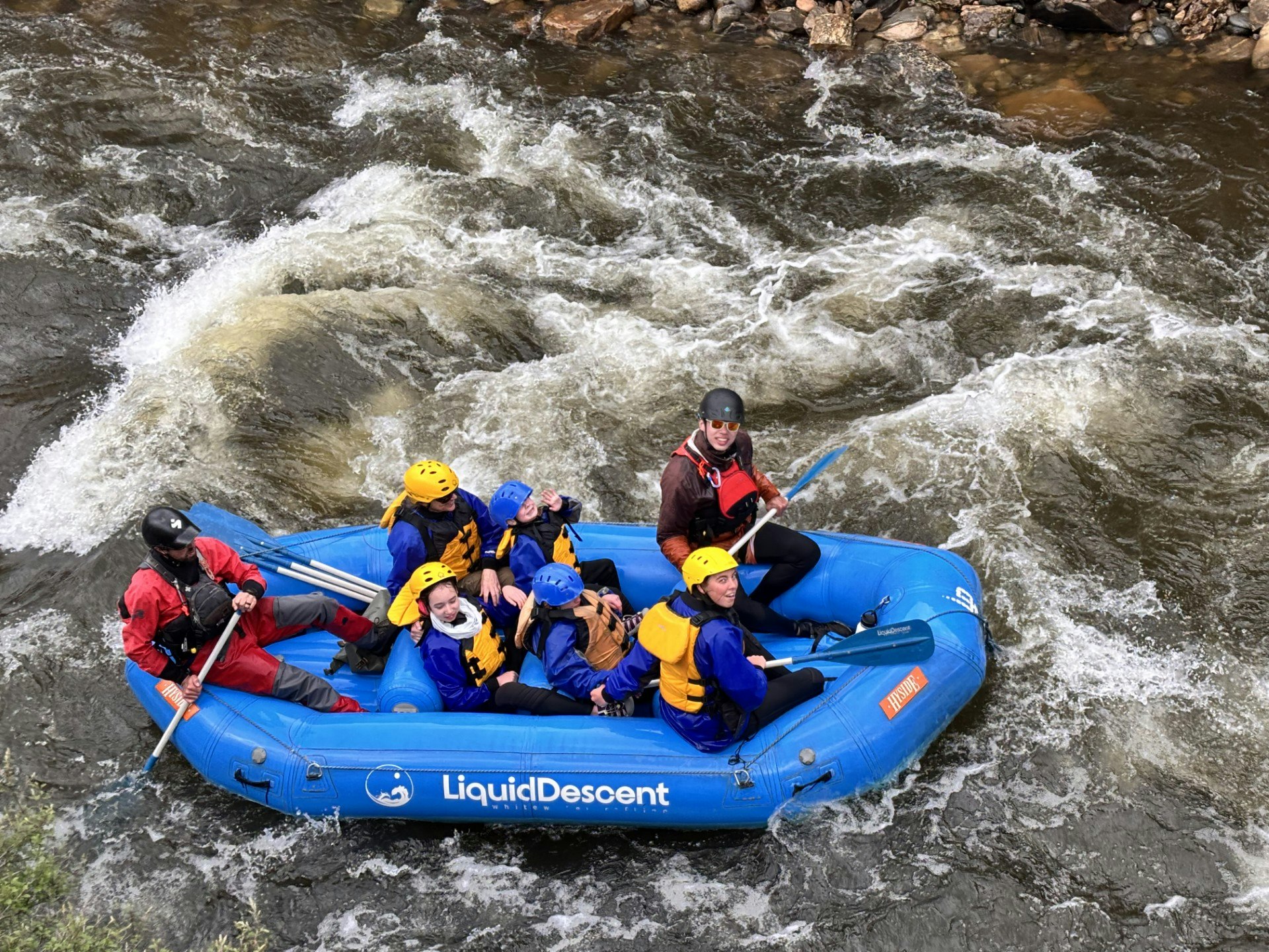 A group of people in helmets and life jackets are white water rafting in a blue raft on a rapid river.