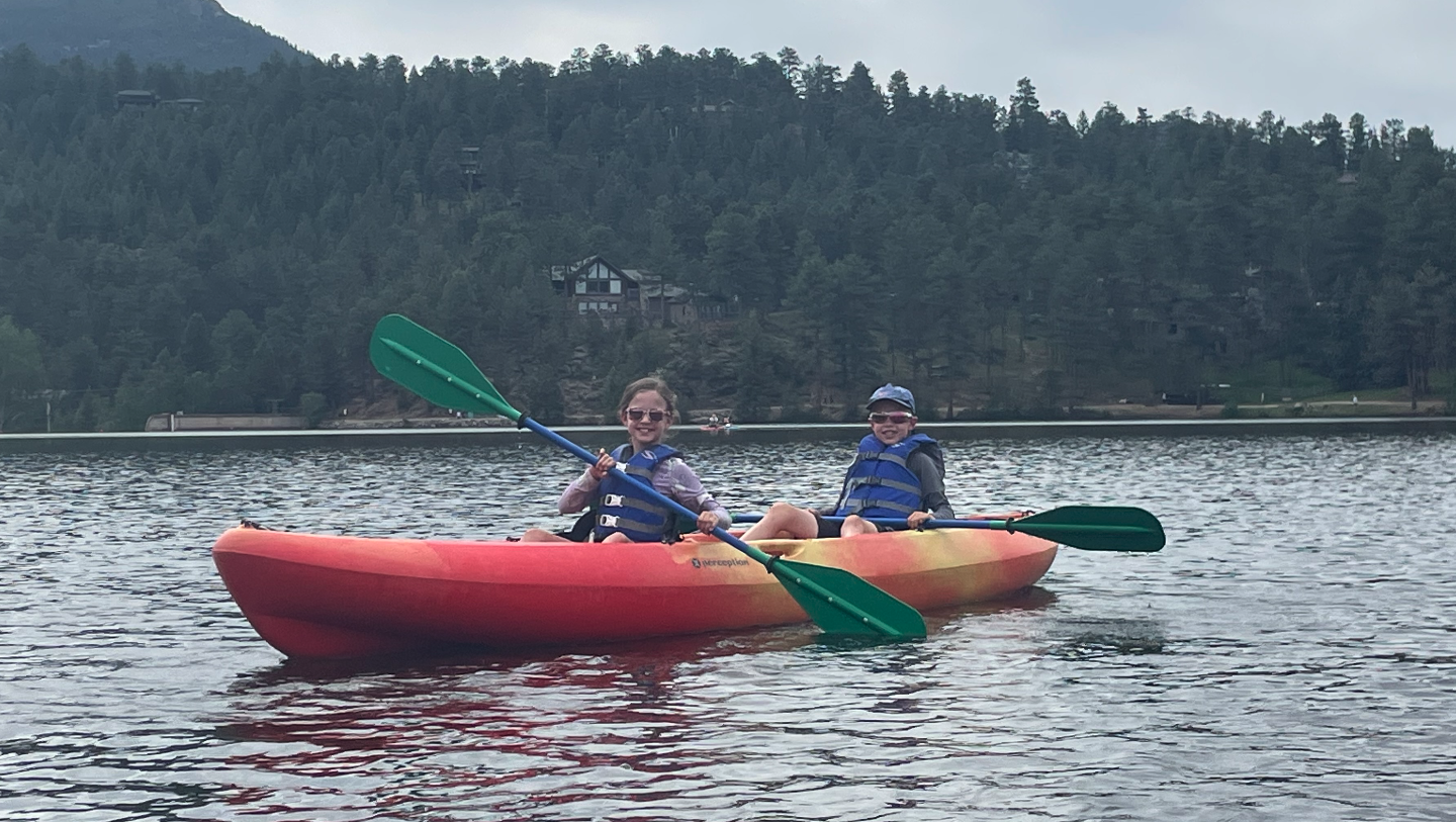 Two people kayaking on a lake with forested hills in the background.