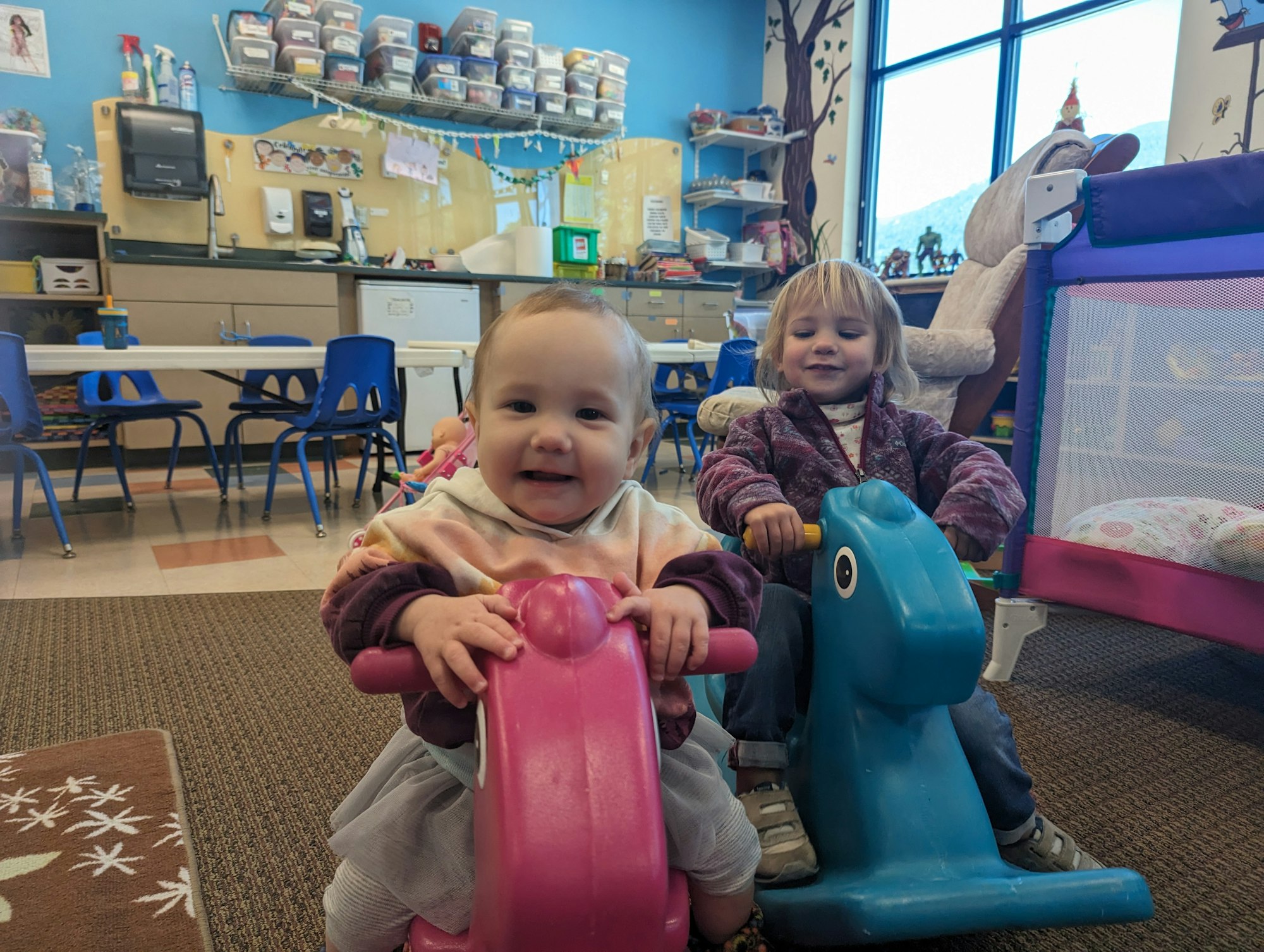 Two children playing on rocking toys in a colorful room with chairs and shelves in the background.