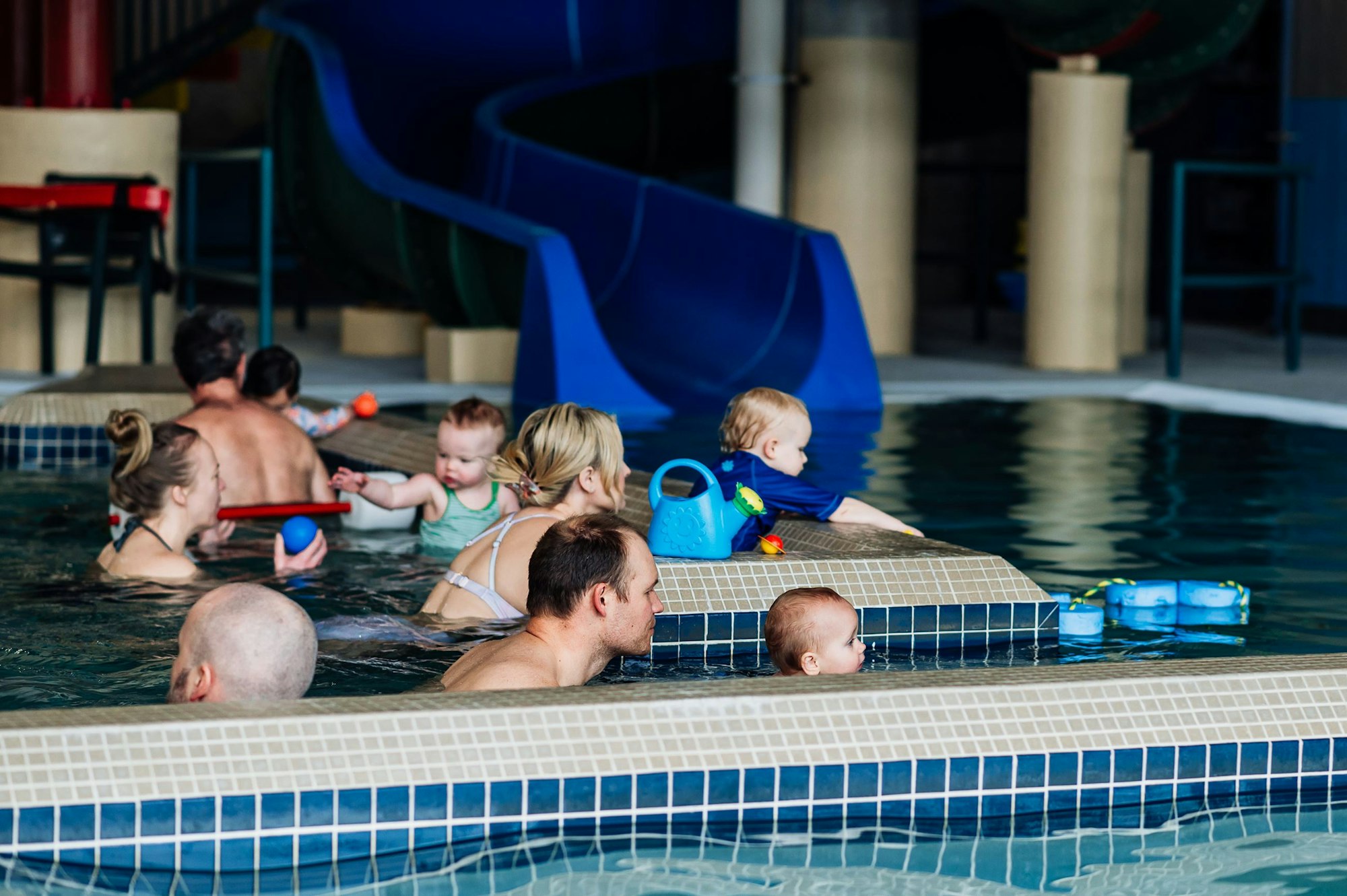 People and children enjoying a pool, with toys and a waterslide in the background.