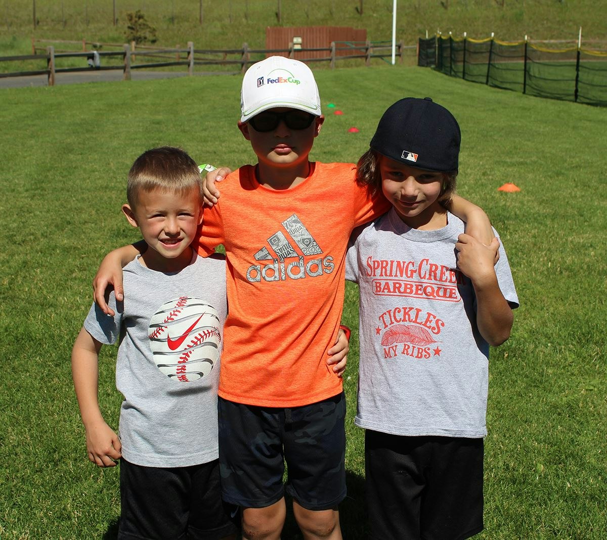 Three kids standing close on a lawn, wearing casual clothes, with arms around each other.