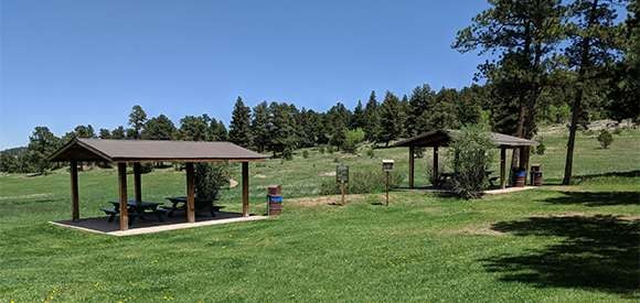 Picnic area with shelters, tables, trash can, and green grass, with trees and hills in the background.