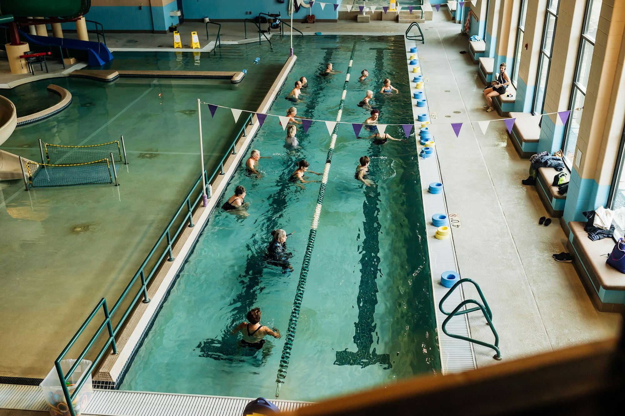 Indoor swimming pool with people in lanes, benches, floats, and a waterslide in the background.