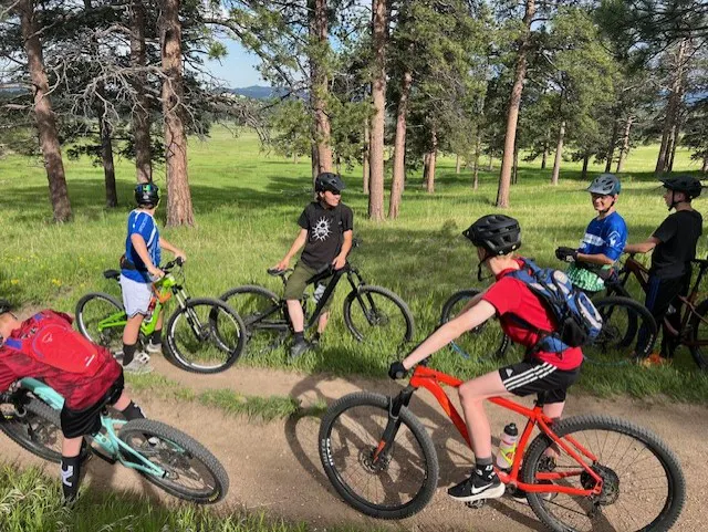 A group of kids on mountain bikes gathers near a trail in a wooded area, enjoying outdoor recreation.
