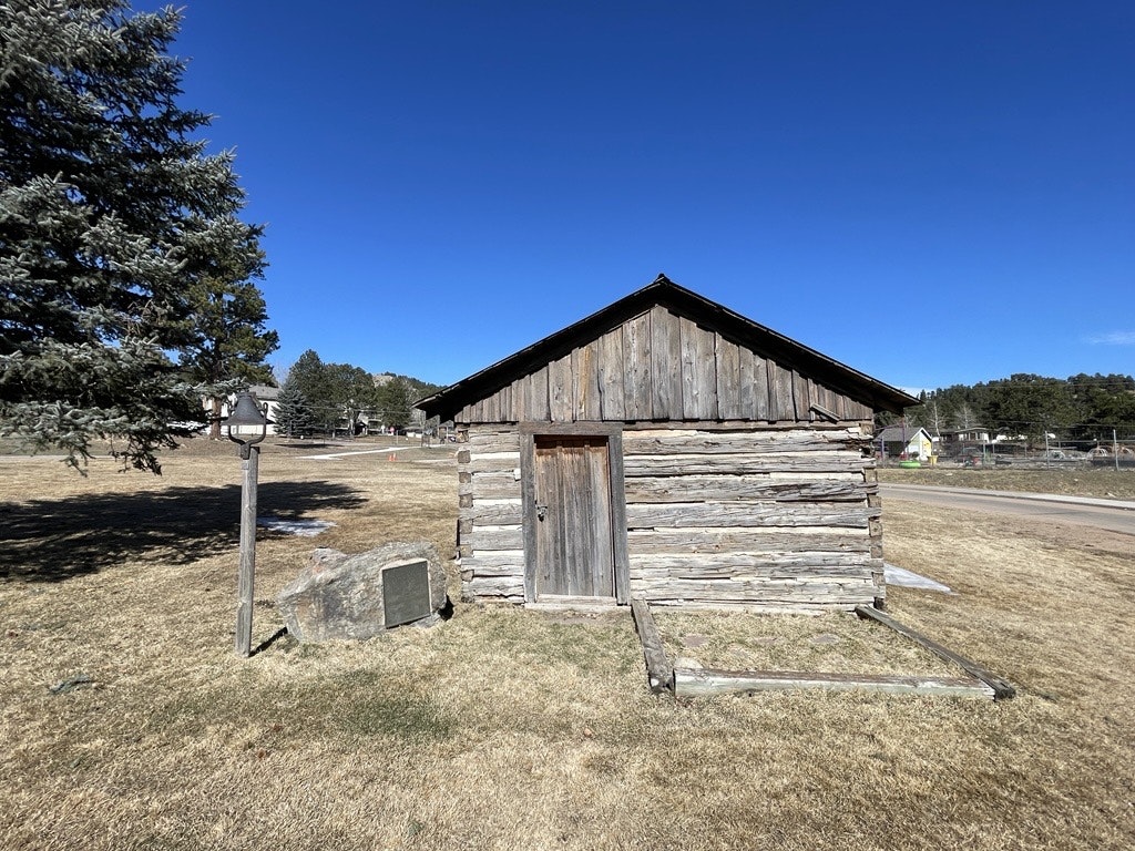 A small, rustic log cabin with a wooden roof and a plaque on a stone nearby, set in a grassy, open field under a clear blue sky.
