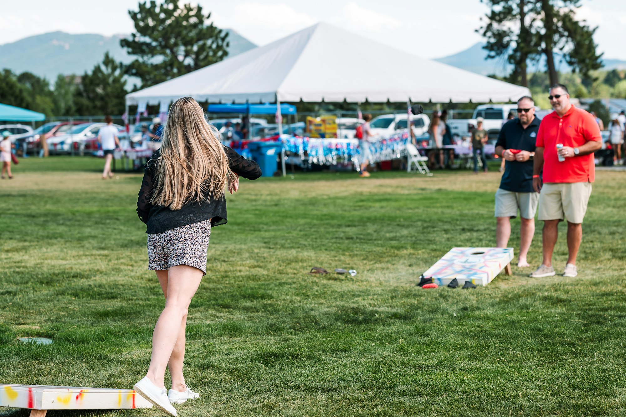 People playing cornhole on a grassy field with tents and trees in the background.
