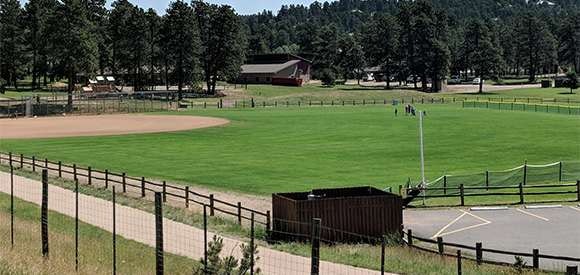 A baseball field, fence, trees, and a red building under a clear sky.