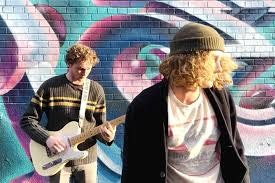 Two individuals are in front of a colorful mural. One plays a guitar while the other turns away, creating a dynamic scene.