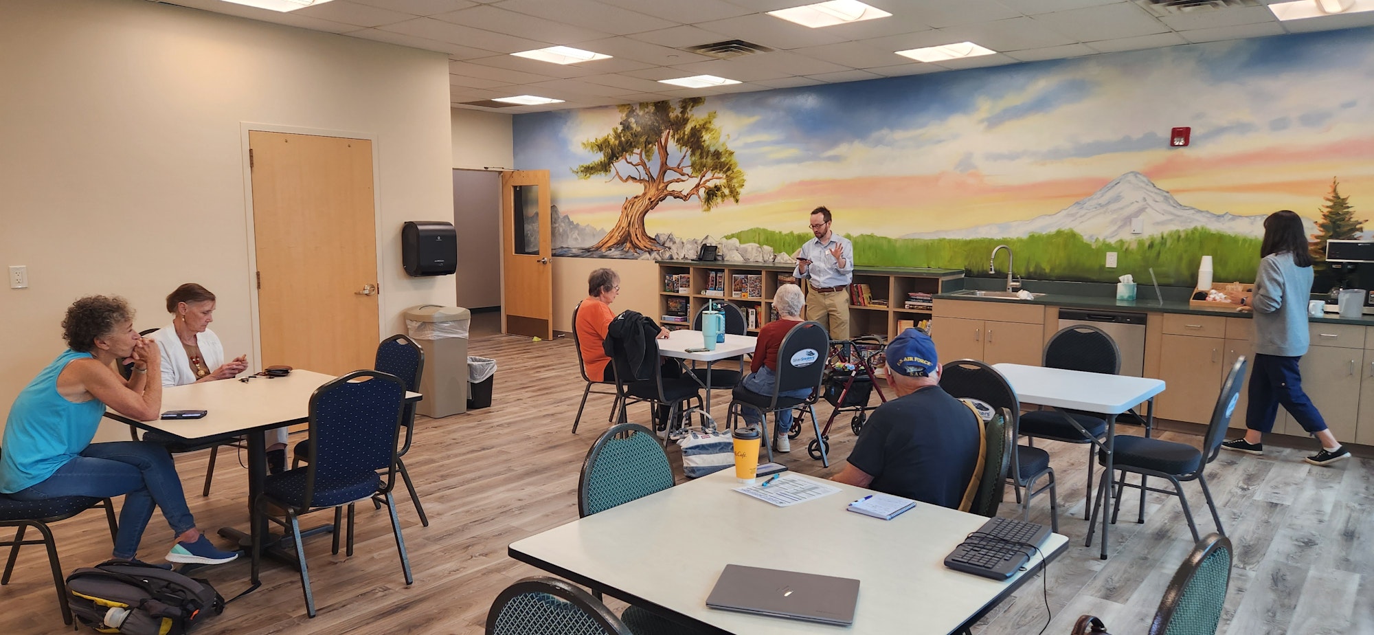 People in a room with tables, chairs, and a mural of mountains and trees. A person is speaking, others are seated or getting coffee.