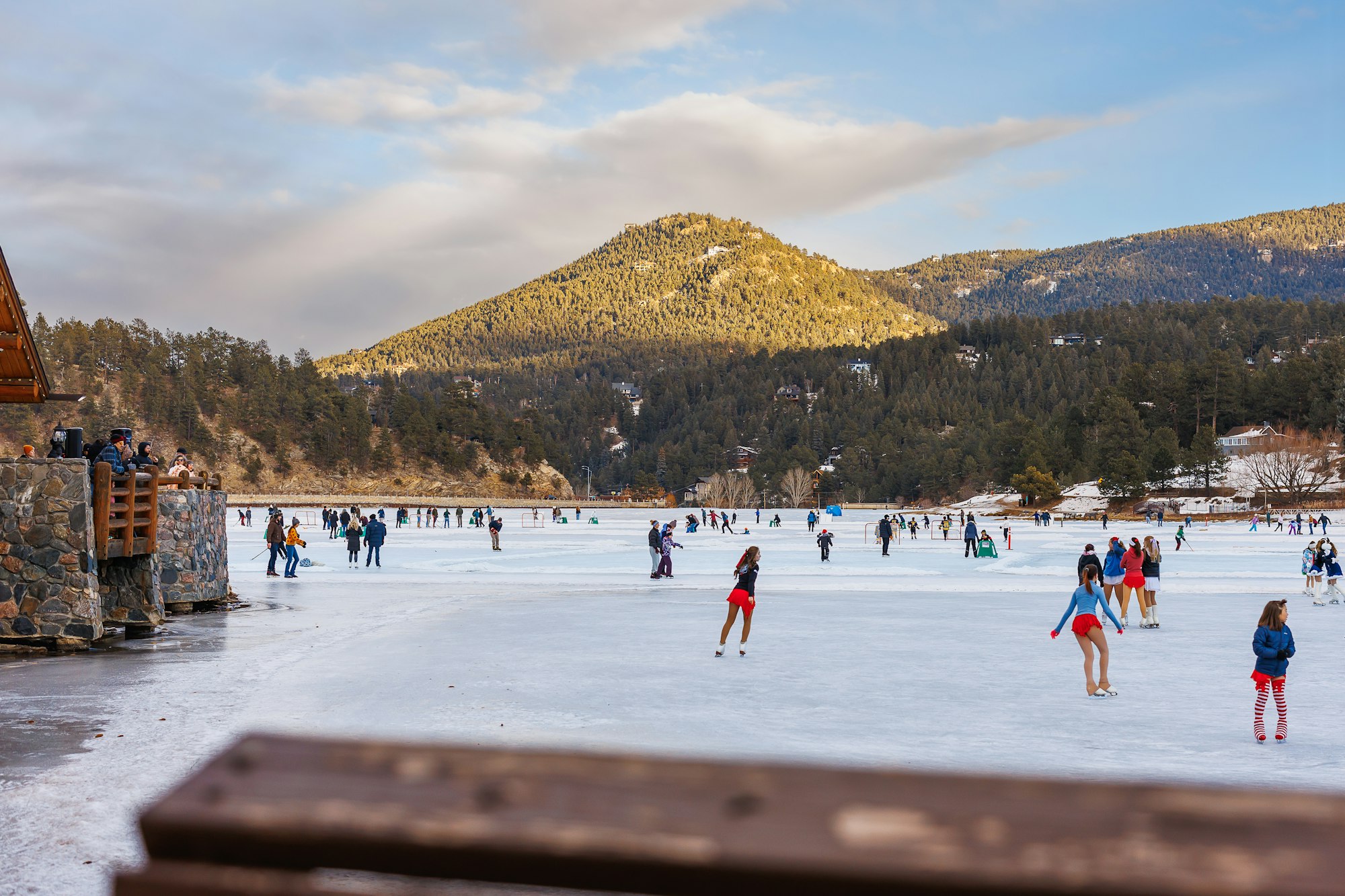 People ice skating on a frozen lake surrounded by mountains and trees under a partly cloudy sky.