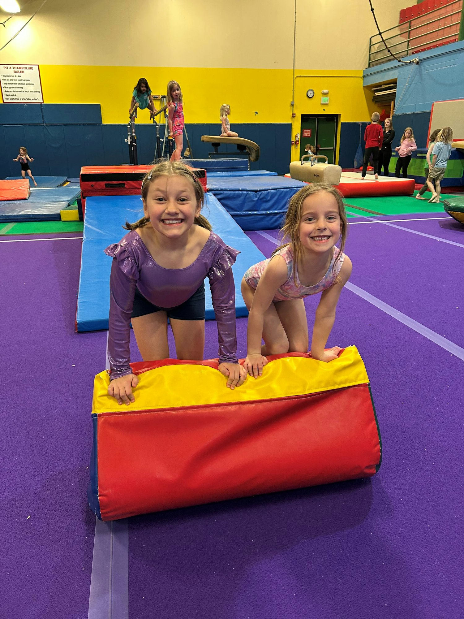 Two smiling girls are playing on colorful gym mats in a gymnastics studio, with other kids and equipment in the background.
