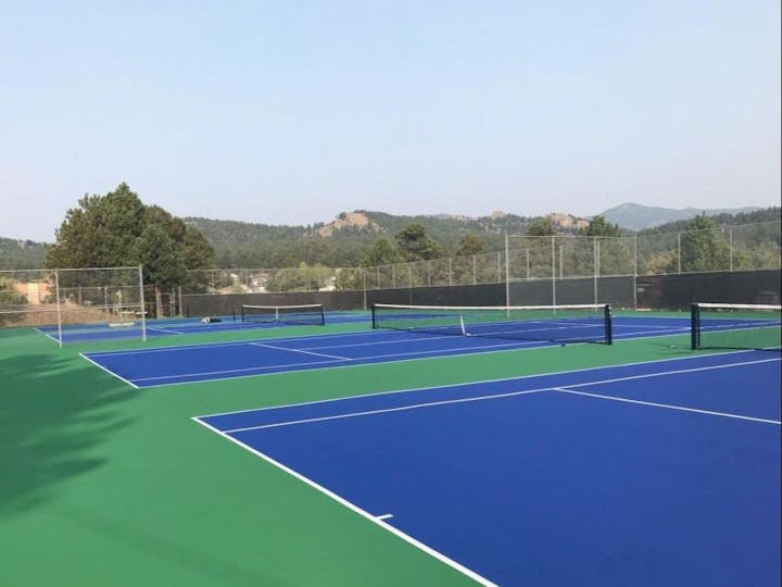 A tennis court with blue and green surface, surrounded by a fence with a hilly landscape in the background.