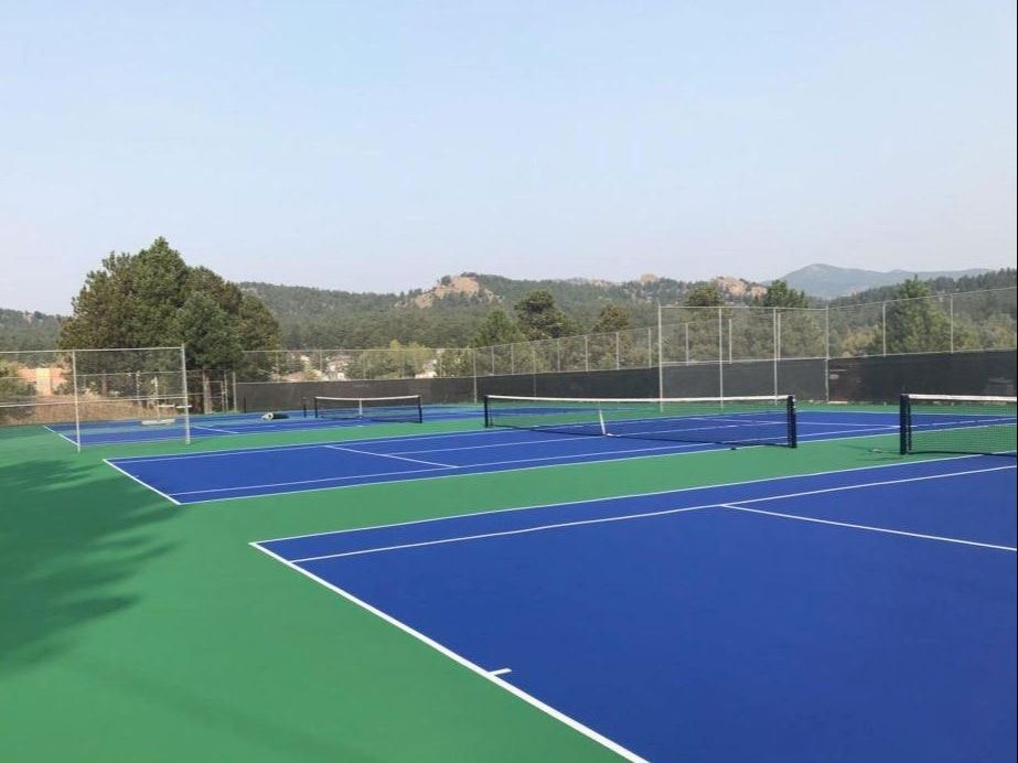 A tennis court with blue and green surface, surrounded by a fence with a hilly landscape in the background.
