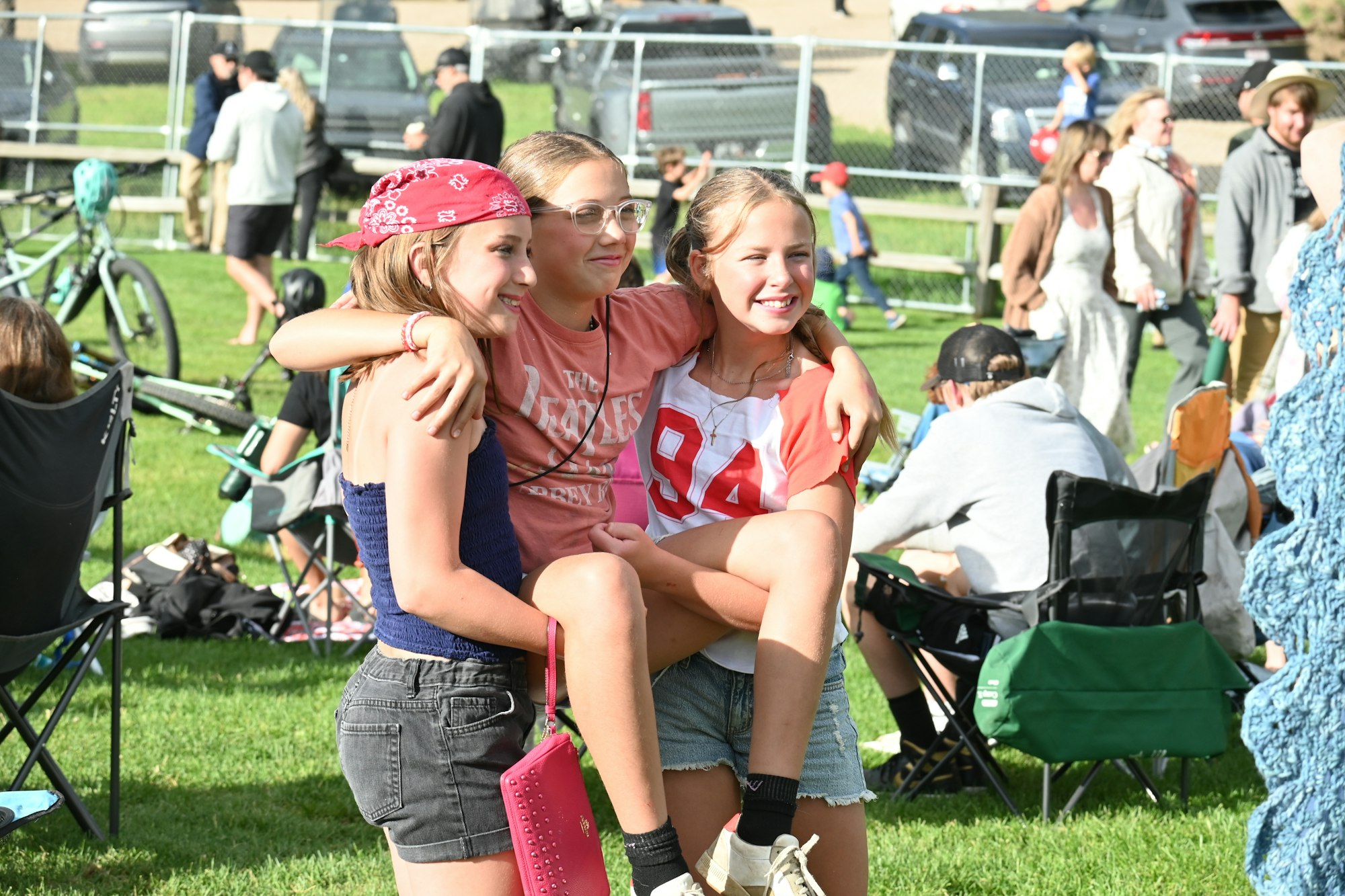 Three people laughing and hugging at an outdoor event, with others relaxing and bicycles in the background.