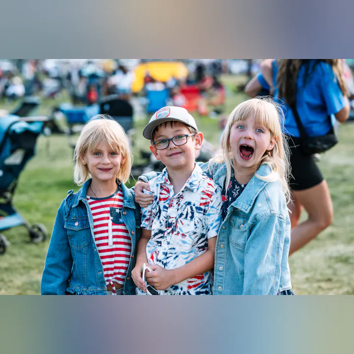 Three children smiling and posing enthusiastically outdoors, dressed in patriotic-themed clothing.