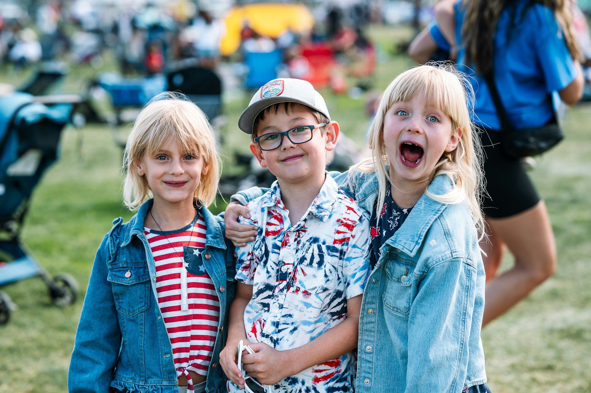 Three children smiling and posing enthusiastically outdoors, dressed in patriotic-themed clothing.