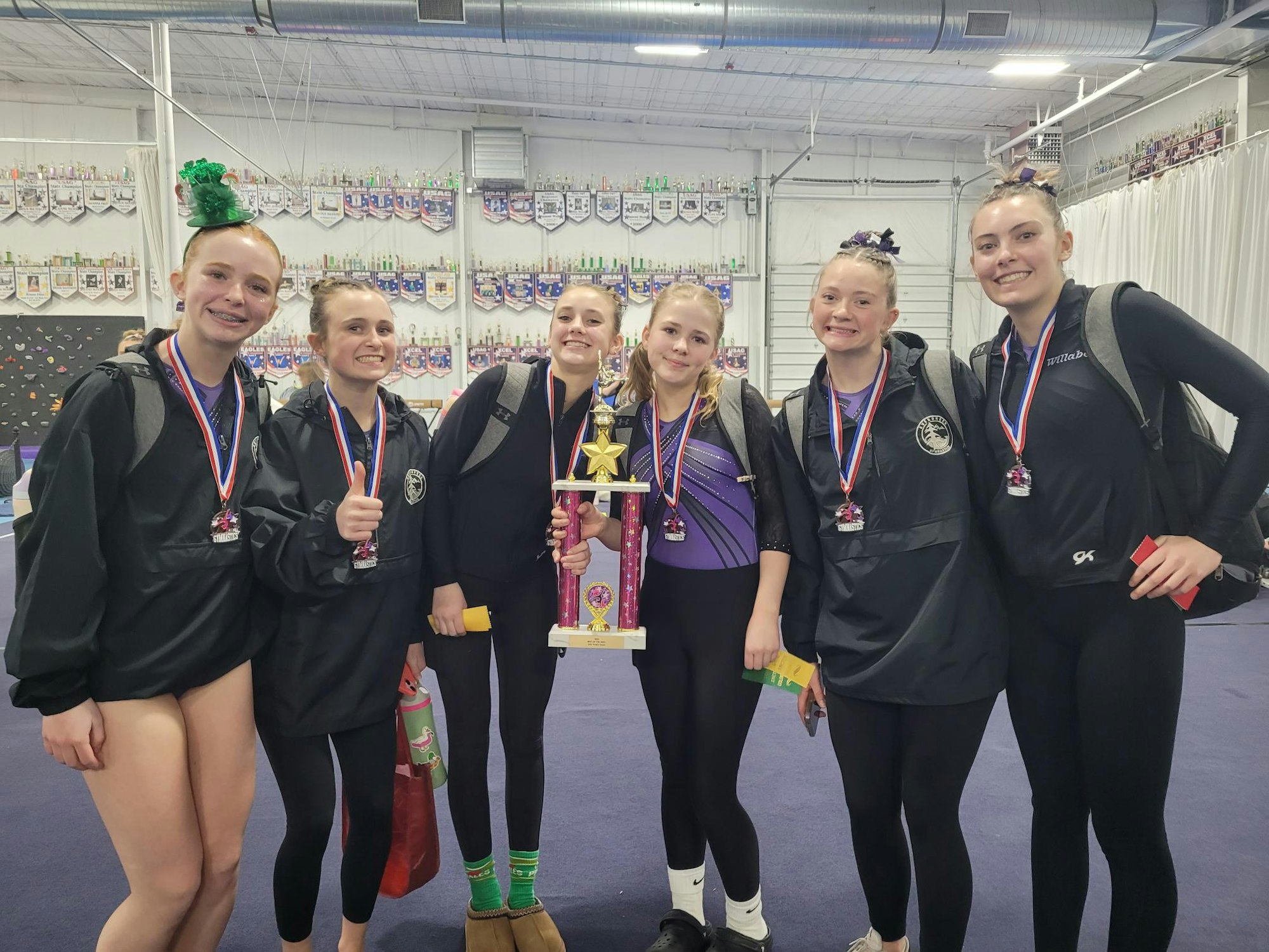Six people wearing medals, holding a trophy, and smiling in a room with banners and trophies in the background.