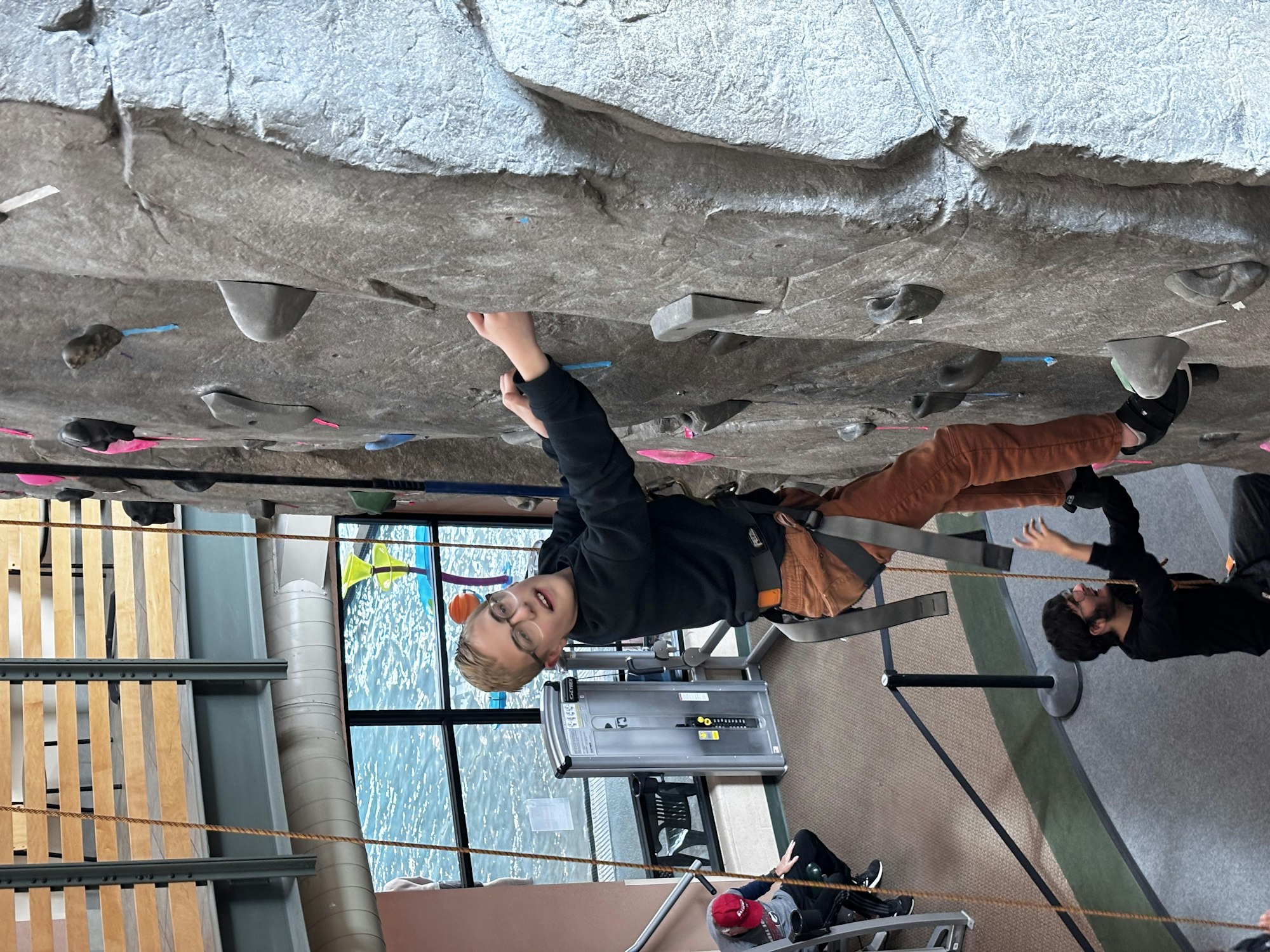 A person is rock climbing indoors on a wall with climbing holds, wearing a harness. Another person is belaying below.