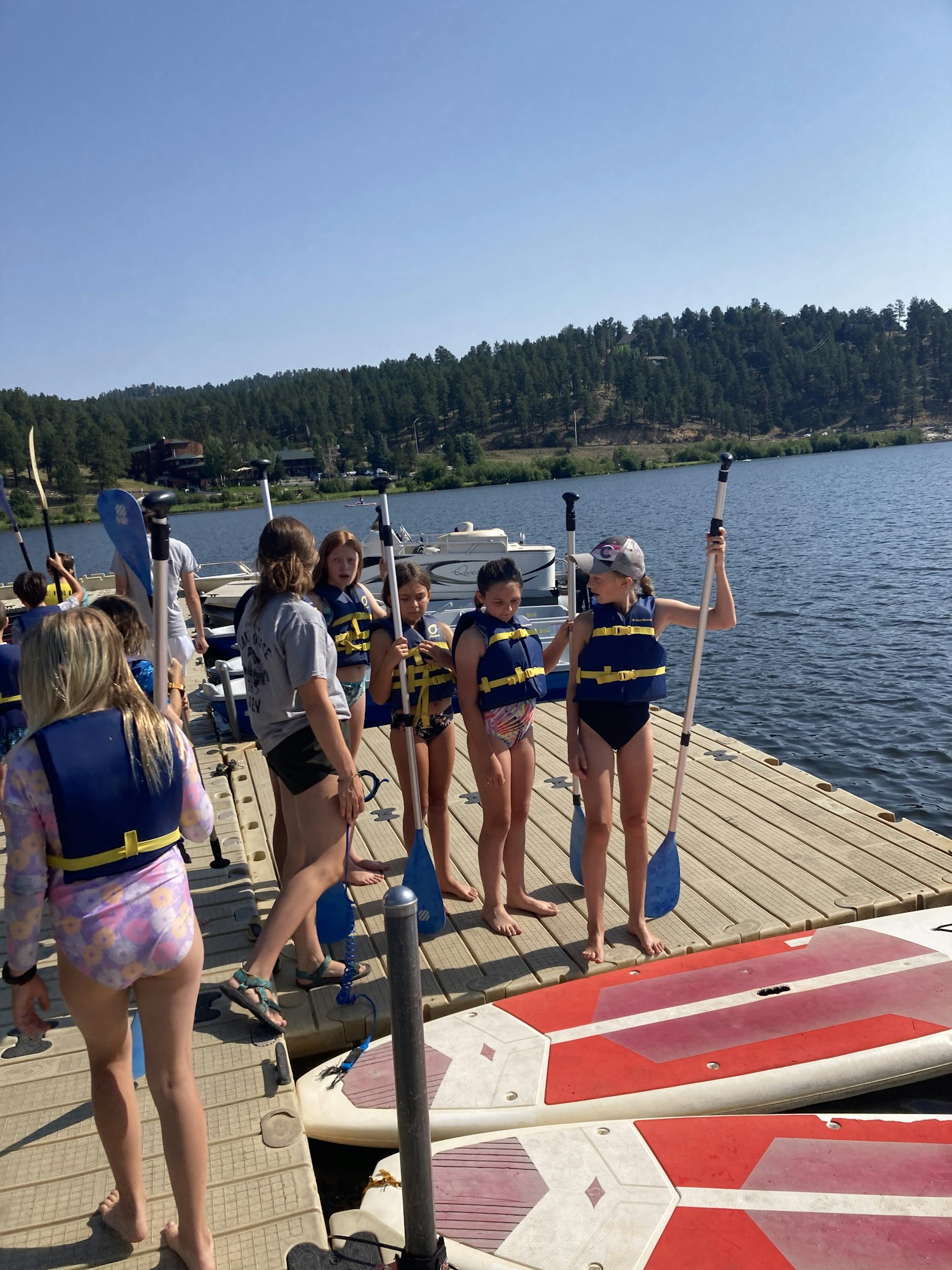A group of people with life jackets prepare for paddleboarding on a dock by the water.