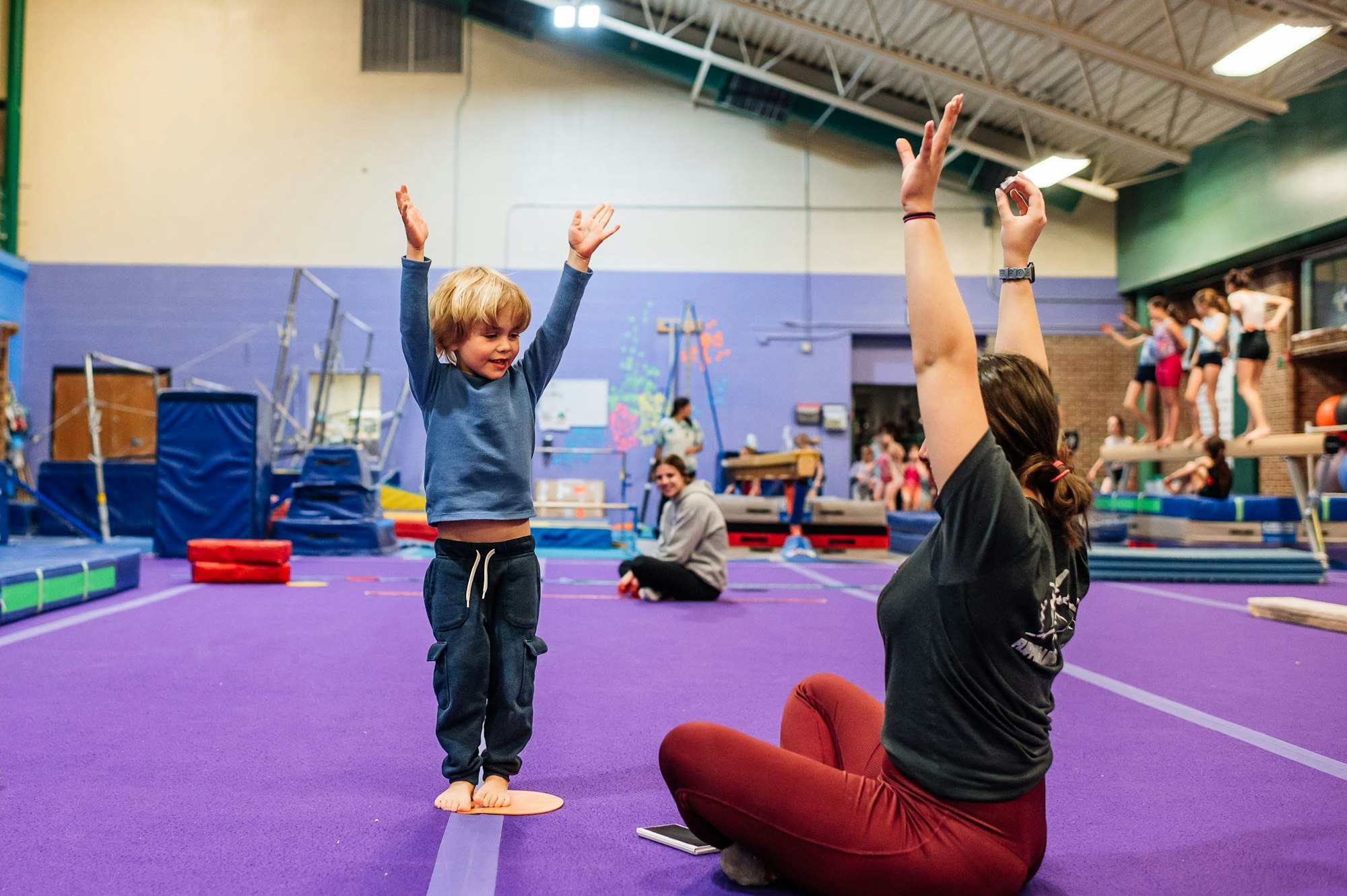 A child and instructor celebrate in a gym with gymnastics equipment and other people in the background.